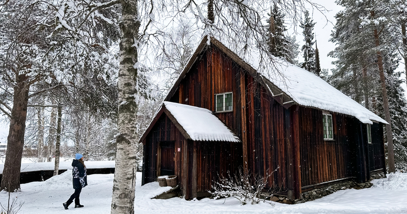 Person walking up to Sodankylä Old Church, a 17th-century wooden church near the Kitinen River in Sodankylä, Lapland. The church has a snow-covered roof and is surrounded by pine trees and winter scenery – peaceful Finnish winter landscape.