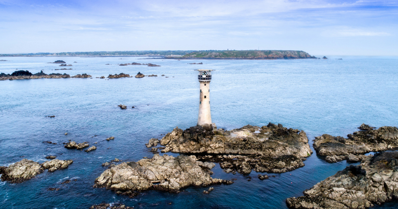 Les Hanois Lighthouse, Guernsey. A tall stone lighthouse on rocky islet surrounded by calm blue sea, with distant coastline and scattered rock formations — remote coastal navigation landmark.