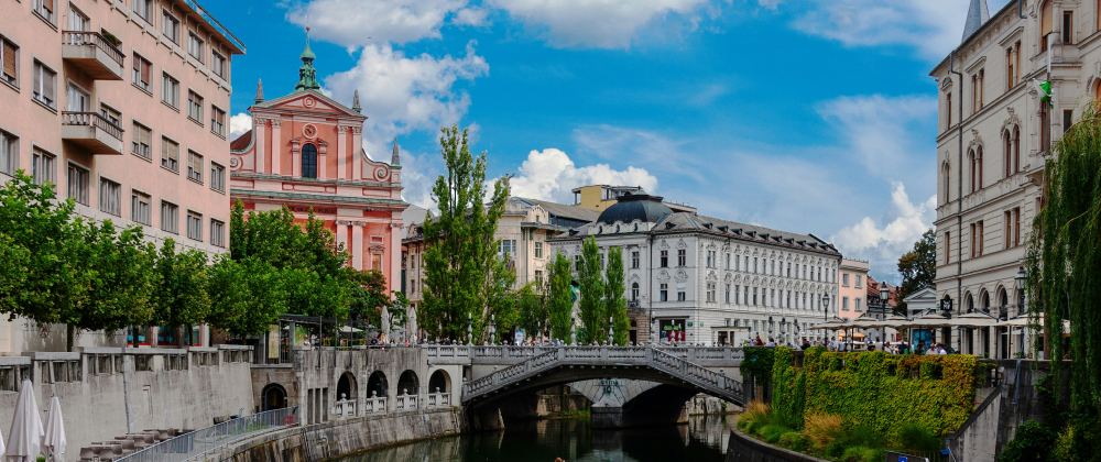 Scenic Ljubljana cityscape with the Ljubljanica River, historic buildings, a stone bridge, leafy trees, and a pink church beneath a blue sky.