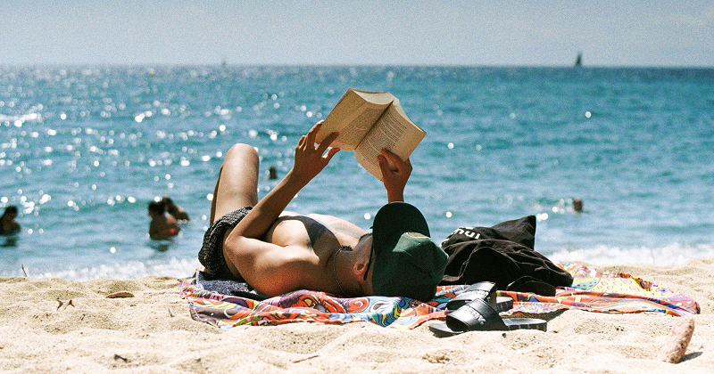 Person lying on a beach towel reading a book under the sun, with waves sparkling in the background and people swimming in the sea – relaxing summer beach read.