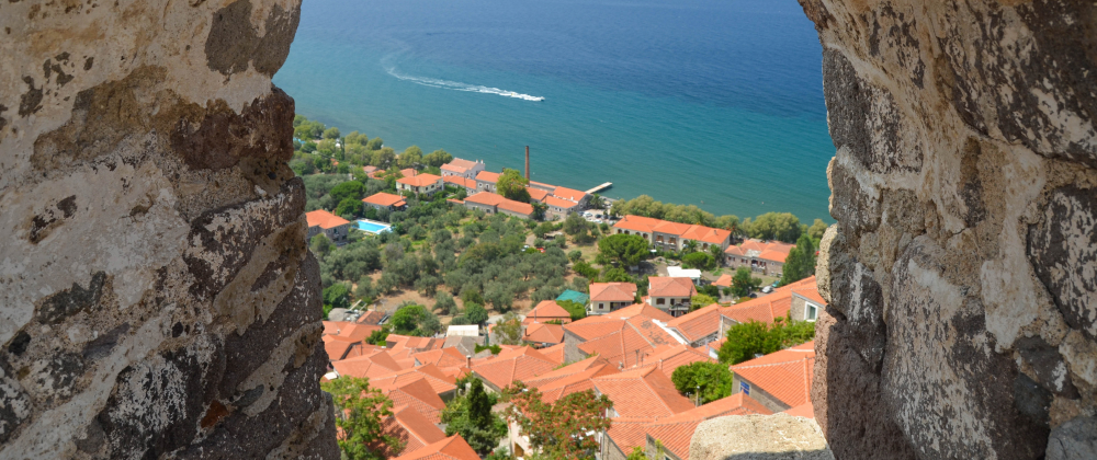 View through stone window of a coastal village with red-tiled roofs, green trees, and blue sea with a passing boat – scenic Mediterranean seaside landscape.