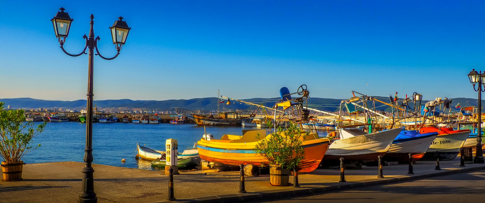 Colourful fishing boats moored in a sunny coastal harbour with calm blue water, lampposts, and distant mountains – peaceful seaside marina scene.