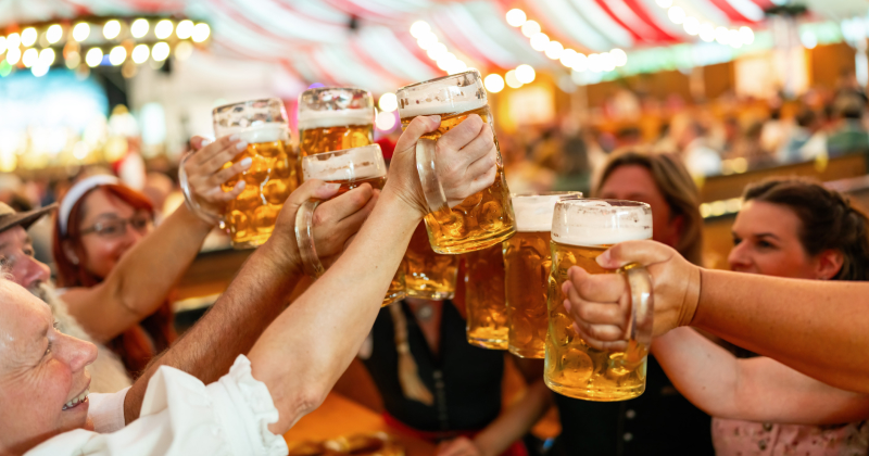 Group of people clinking large beer mugs at a lively Oktoberfest beer tent with red and white striped canopy and festive lights.