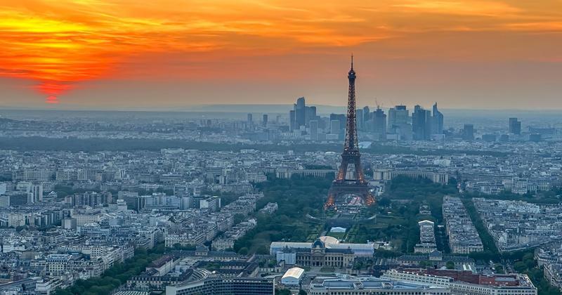 Eiffel Tower rising above Paris cityscape at sunset, with orange sky and modern skyline in the distance – romantic evening view of Paris.