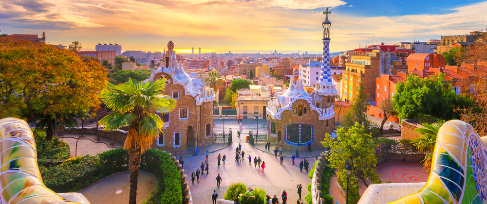Tourists walking through colourful entrance of Park Güell in Barcelona at sunset, with Gaudí's mosaic buildings, palm trees, and city skyline in view.