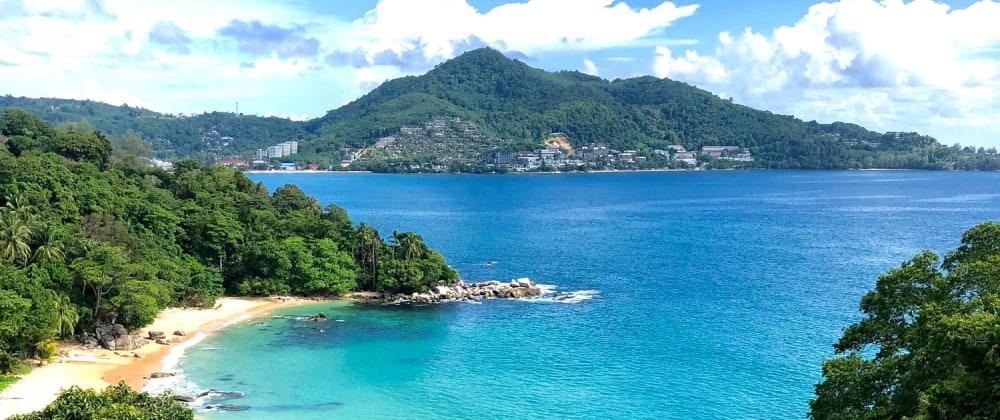 Tropical beach with golden sand and turquoise water, surrounded by lush green forest and overlooking a coastal town on a forested mountain – serene island scenery in Phuket, Thailand.