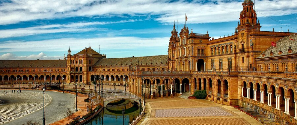 Plaza de España in Seville, Spain, featuring ornate historic buildings, arched colonnades, a canal with a bridge, and a wide tiled plaza under blue sky.