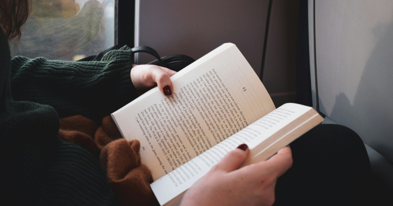 Person reading a paperback book, cosy in a green sweater with brown scarf, natural light through window – quiet travel moment.