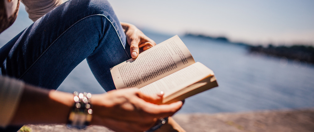 Person reading a paperback book by a calm lake, wearing jeans and bracelets – peaceful outdoor relaxation with scenic waterfront view.