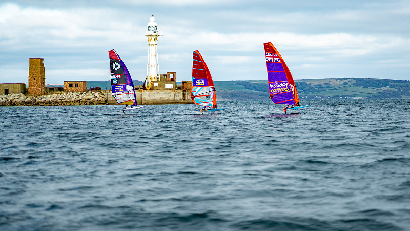 Three windsurfers racing on open water near a coastal lighthouse, colorful sails raised under a cloudy sky during competitive windsurfing.