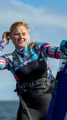 Female windsurfer smiling on the water, wearing a colorful wetsuit and harness, holding her sail in windy conditions under a blue sky.