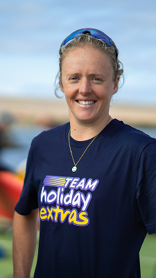 Smiling woman wearing a Team Holiday Extras T-shirt, standing outdoors with sports sunglasses on her head at a windsurfing event.