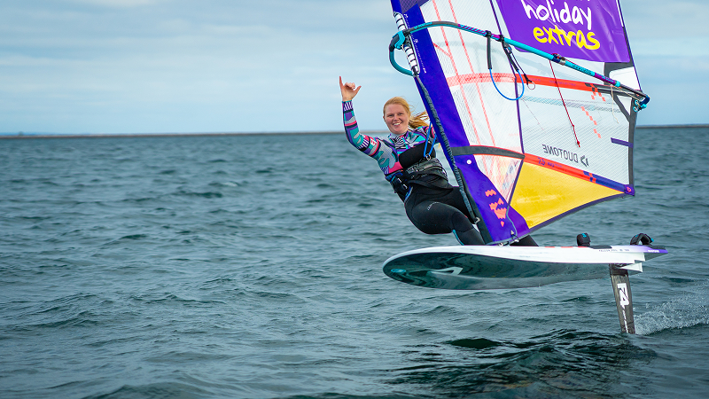 Smiling female windsurfer riding a hydrofoil board, lifting one hand while gliding above the water with a colorful sail.