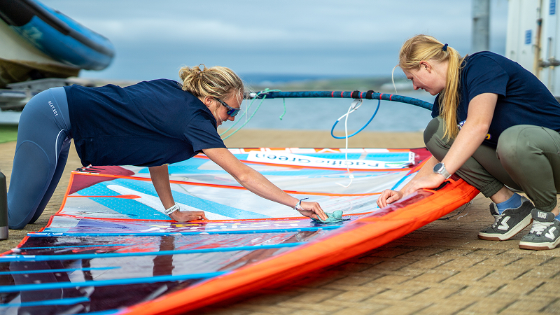 Two women preparing a windsurf sail on the ground, adjusting lines and battens together before heading out on the water.