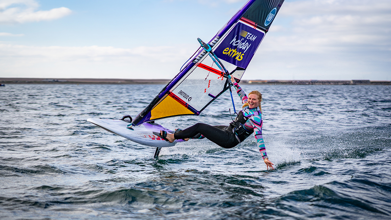 Female windsurfer performing a fast hydrofoil maneuver, leaning low with one hand touching the water during high-speed sailing.