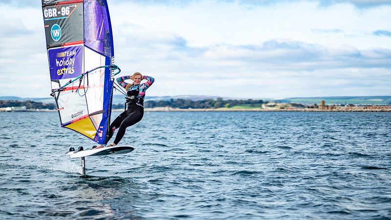 Female windsurfer riding a hydrofoil board, lifting both hands while gliding above the water with a colorful sail on a coastal bay.