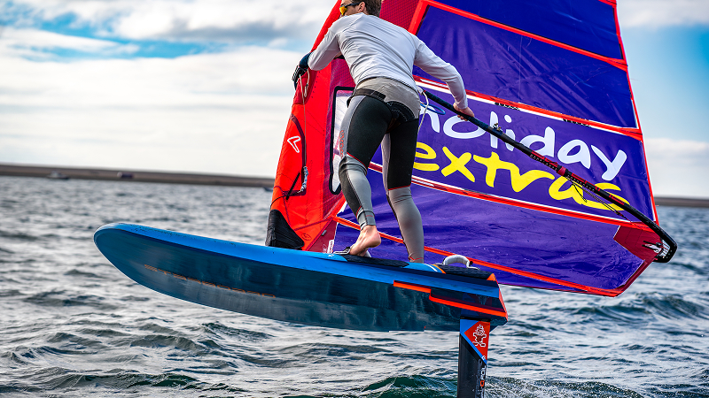 Windsurfer viewed from behind balancing on a hydrofoil board, colorful sail powered up while flying above choppy water.
