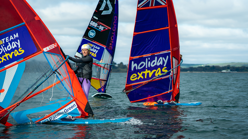 Group of windsurfers standing on boards in shallow water, holding colorful racing sails during a training session on a coastal bay.