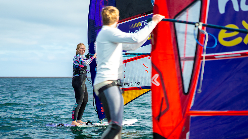 Two windsurfers maneuver past each other on calm water, colorful sails close together during a windsurfing practice session.