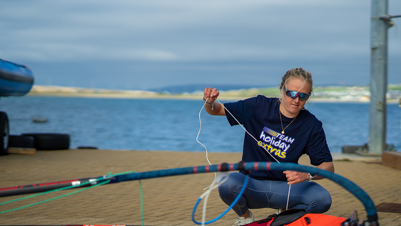 Female windsurfer crouching on shore, threading lines while rigging a windsurf sail beside the water under cloudy skies.