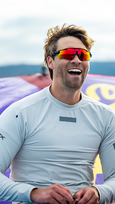 Smiling windsurfer wearing red mirrored sports sunglasses and a light grey performance top, standing outdoors with a colorful sail behind him.