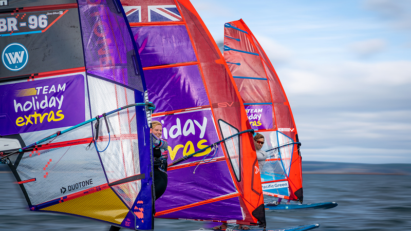 Close-up of windsurfers racing side by side on hydrofoil boards, colorful sails cutting through choppy water at speed.