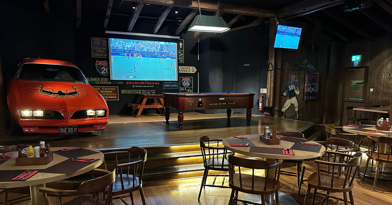 Sheraton Skyline sports bar interior with a classic red muscle car display, large screens showing a soccer match, pool table stage, wooden tables, and vintage Americana decor.
