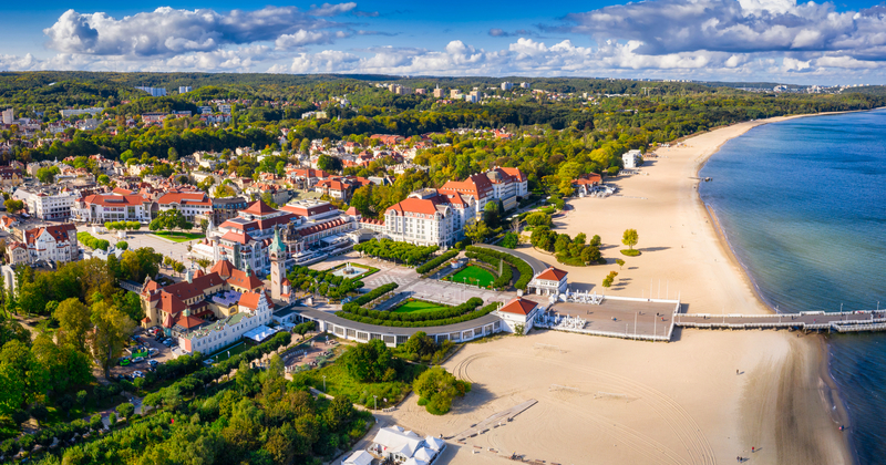 Aerial view of a coastal resort town with a wide sandy beach, seaside hotels, lush green parks, and a long pier along the Baltic Sea under a bright blue sky.