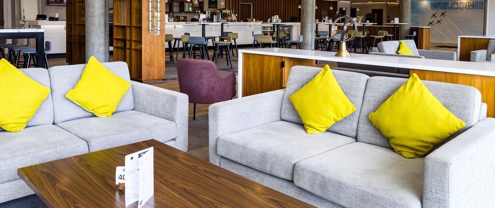 Modern hotel lounge at Hampton by Hilton Stansted with grey sofas, bright yellow cushions, wooden coffee tables, and bar seating in the background.