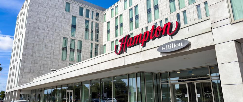Exterior view of Hampton by Hilton hotel at Stansted Airport, with modern grey facade, large windows, and bold red signage above glass entrance.