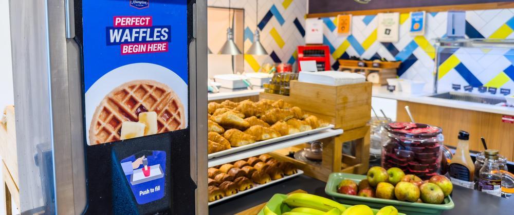 Self-serve waffle station with pastries, apples, bananas, and syrups at Hampton by Hilton Stansted Airport breakfast buffet.