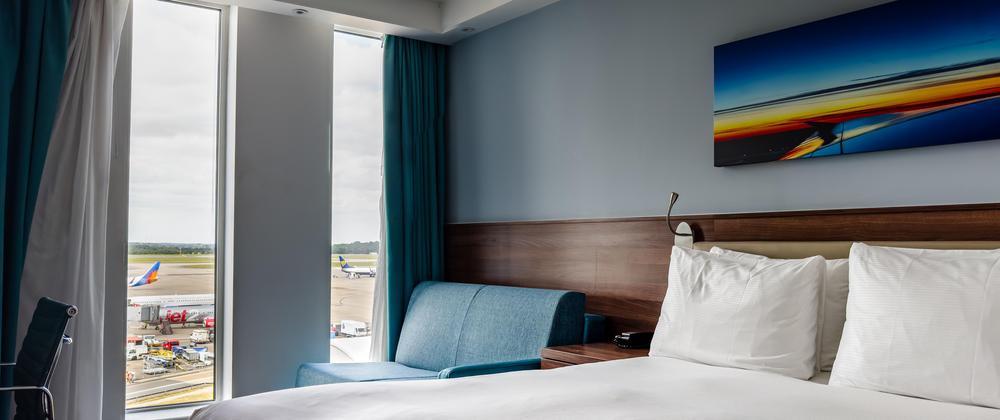 Airport-facing guest room at Hampton by Hilton Stansted with king bed, blue sofa, modern decor, and runway views through tall windows.