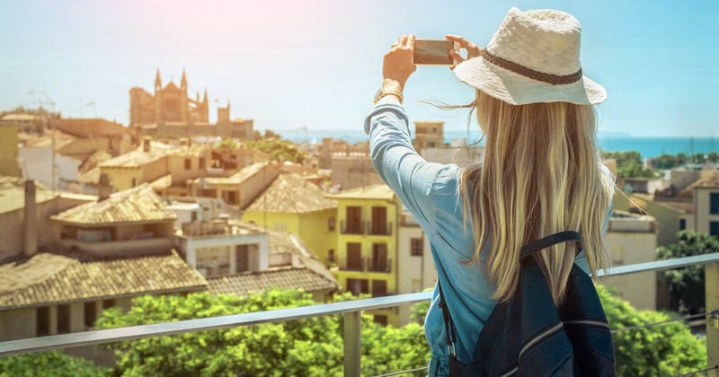 Woman with long blonde hair taking a photo of a sunny Mediterranean cityscape with historic buildings, cathedral, and sea in the distance.
