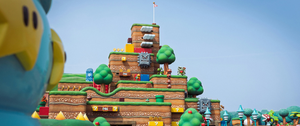 Colourful Super Mario platform game scene with grassy hills, trees, coins, pipes, Thwomps, enemies, and layered obstacle-filled terrain. Super Nintendo World, Japan.