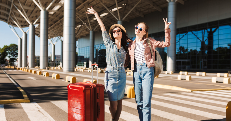 Two happy young women with luggage posing outside airport terminal, wearing sunglasses and casual outfits — excited travel departure moment.
