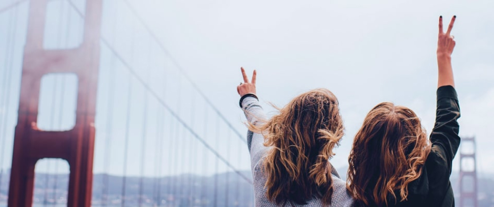Two women with windblown hair raising peace signs on the Golden Gate Bridge, overlooking San Francisco Bay – joyful travel moment in California.