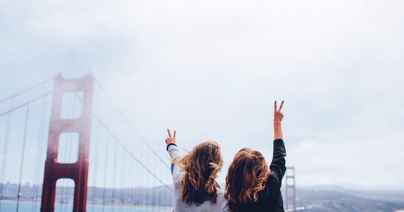 Two women with windblown hair raising peace signs on the Golden Gate Bridge, overlooking San Francisco Bay – joyful travel moment in California.