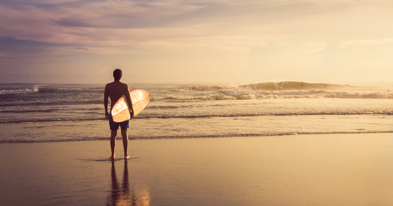 Surfer standing on wet sand holding a board, gazing at gentle waves during golden sunset — peaceful beach scene and surf travel inspiration.