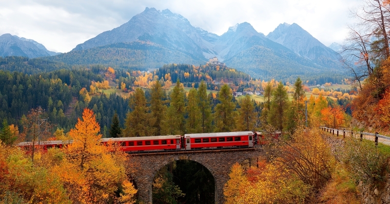The Glacier Express, a red passenger train crossing a stone viaduct through colorful autumn forest with alpine mountains in the background — scenic autumnal travel in the Swiss Alps.