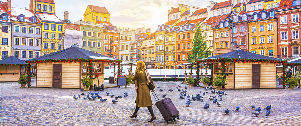 Woman with rolling luggage walking through a festive Christmas market in Warsaw's Old Town Square, surrounded by pigeons and colourful historic buildings.