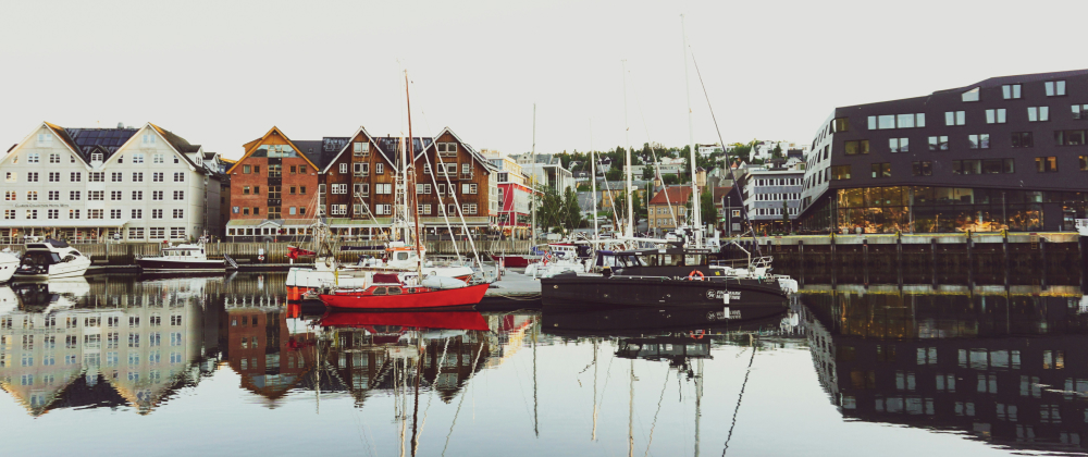 Colourful wooden buildings and modern black architecture reflect in calm harbor waters with moored sailboats in Tromsø, Norway — scenic waterfront cityscape.