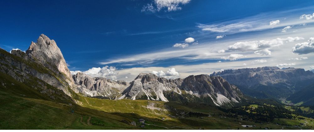 Panoramic view of the Seceda mountain range in Italy's Dolomites beneath a blue sky with scattered clouds.