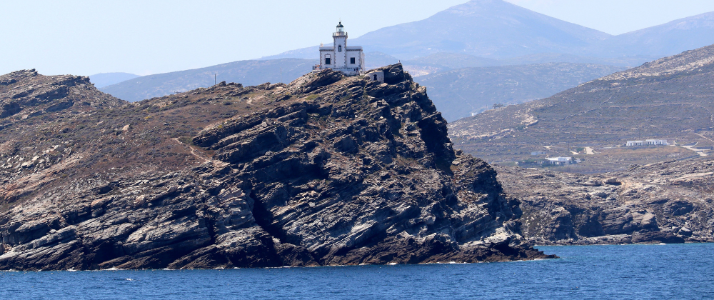 A white lighthouse sits atop a rugged rocky headland overlooking deep blue sea, with hazy mountains and a dry coastal landscape stretching into the distance.