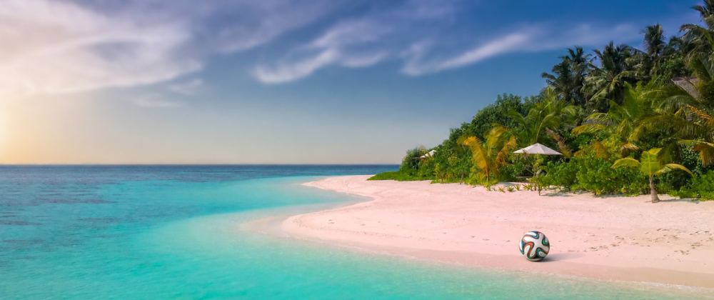 A tropical beach with pale pink-white sand curves along clear turquoise water. Dense palm trees and lush green vegetation line the shore under a blue sky with wispy clouds, while a football rests near the water’s edge.