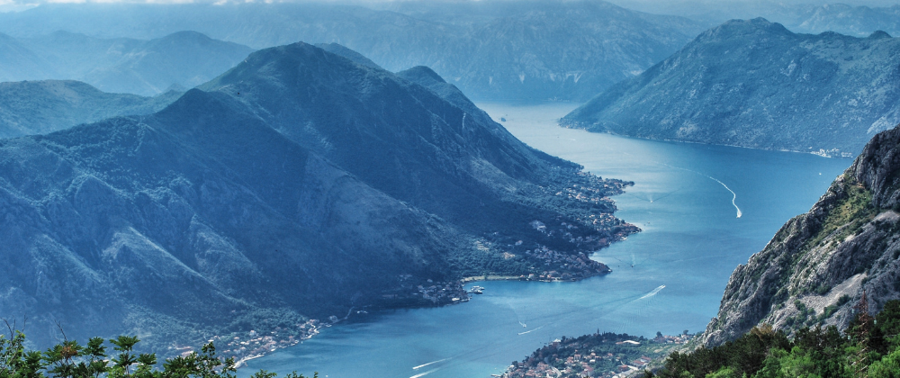 A sweeping view of a winding bay surrounded by steep mountains, with small coastal settlements dotted along the shoreline and boats leaving trails across the water.