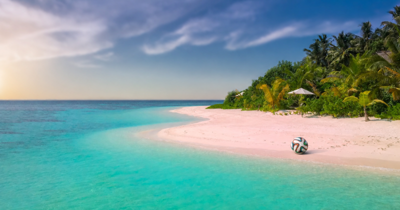A tropical beach with pale pink-white sand curves along clear turquoise water. Dense palm trees and lush green vegetation line the shore under a blue sky with wispy clouds, while a football rests near the water’s edge.