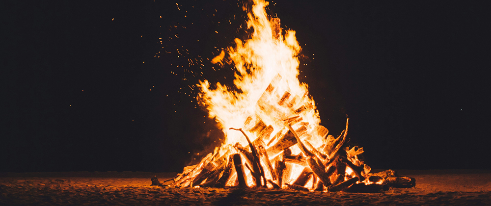 Large bonfire burning brightly at night with stacked logs and glowing embers on a sandy surface — dramatic outdoor fire under a dark sky.