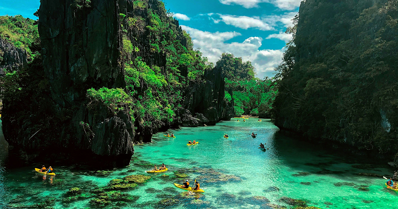 Kayakers paddling through a clear turquoise lagoon surrounded by towering limestone cliffs and lush tropical forest – serene island nature adventure.