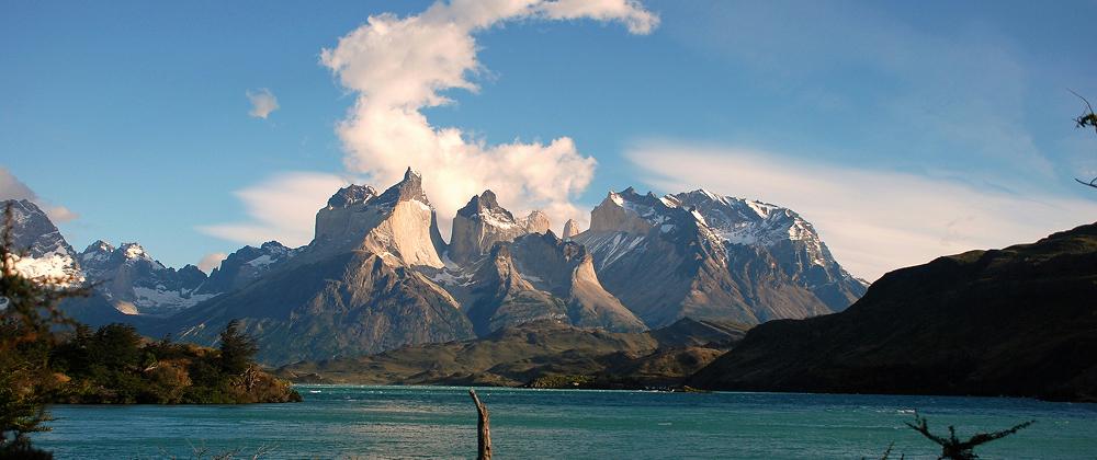 Snow-capped peaks of Torres del Paine rising above a turquoise lake in Patagonia, Chile — dramatic mountain landscape under a bright blue sky.