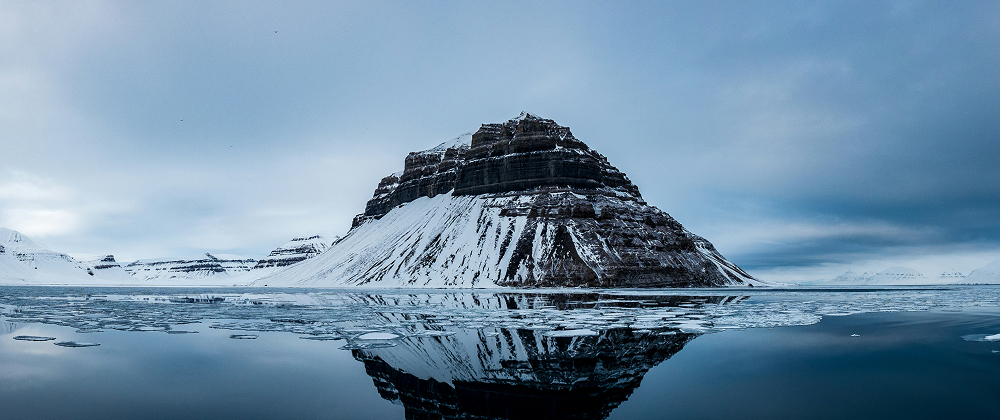 Snow-covered mountain reflected in icy Arctic waters under a cloudy blue sky — remote and dramatic polar landscape.